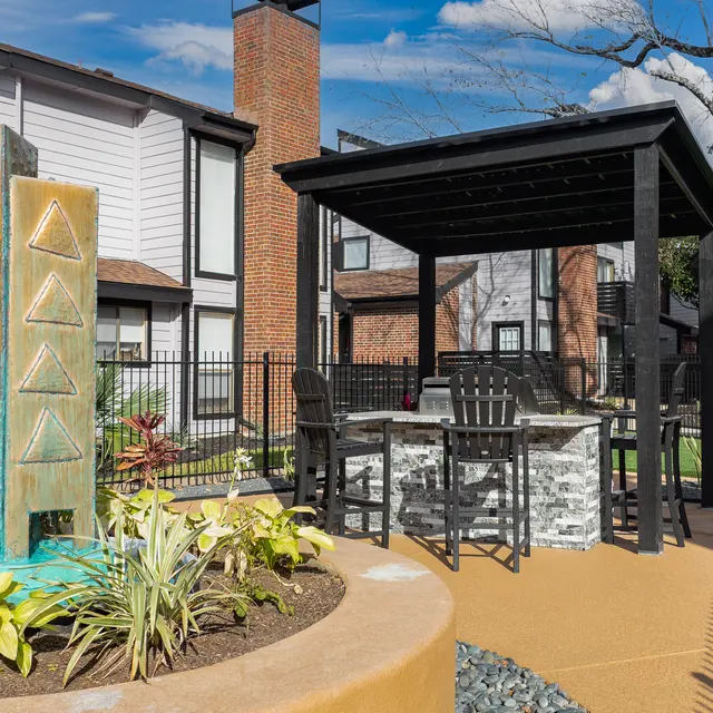 A landscaped area in an apartment complex featuring a modern stone water feature, a gazebo with dining furniture, and surrounding greenery.