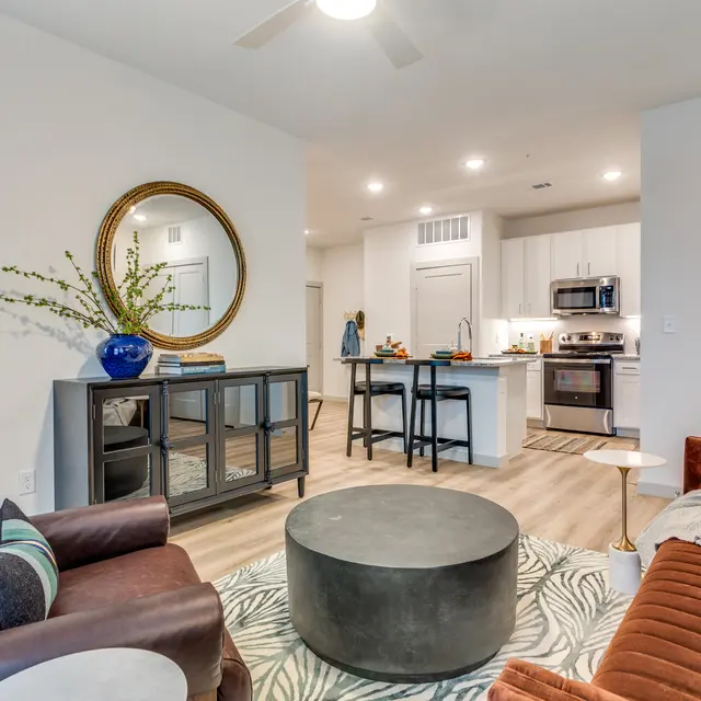 A modern apartment living space featuring brown and orange sofas, a round coffee table, a kitchen view in the background, and decorative elements like a large round mirror and greenery.