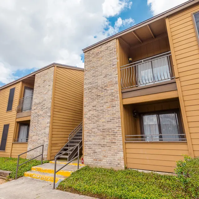 View of an apartment complex with brown siding, brick accents, and a green landscaped area.