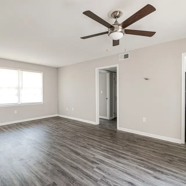 Spacious room with light-colored walls, wooden flooring, a ceiling fan, and windows providing natural light.