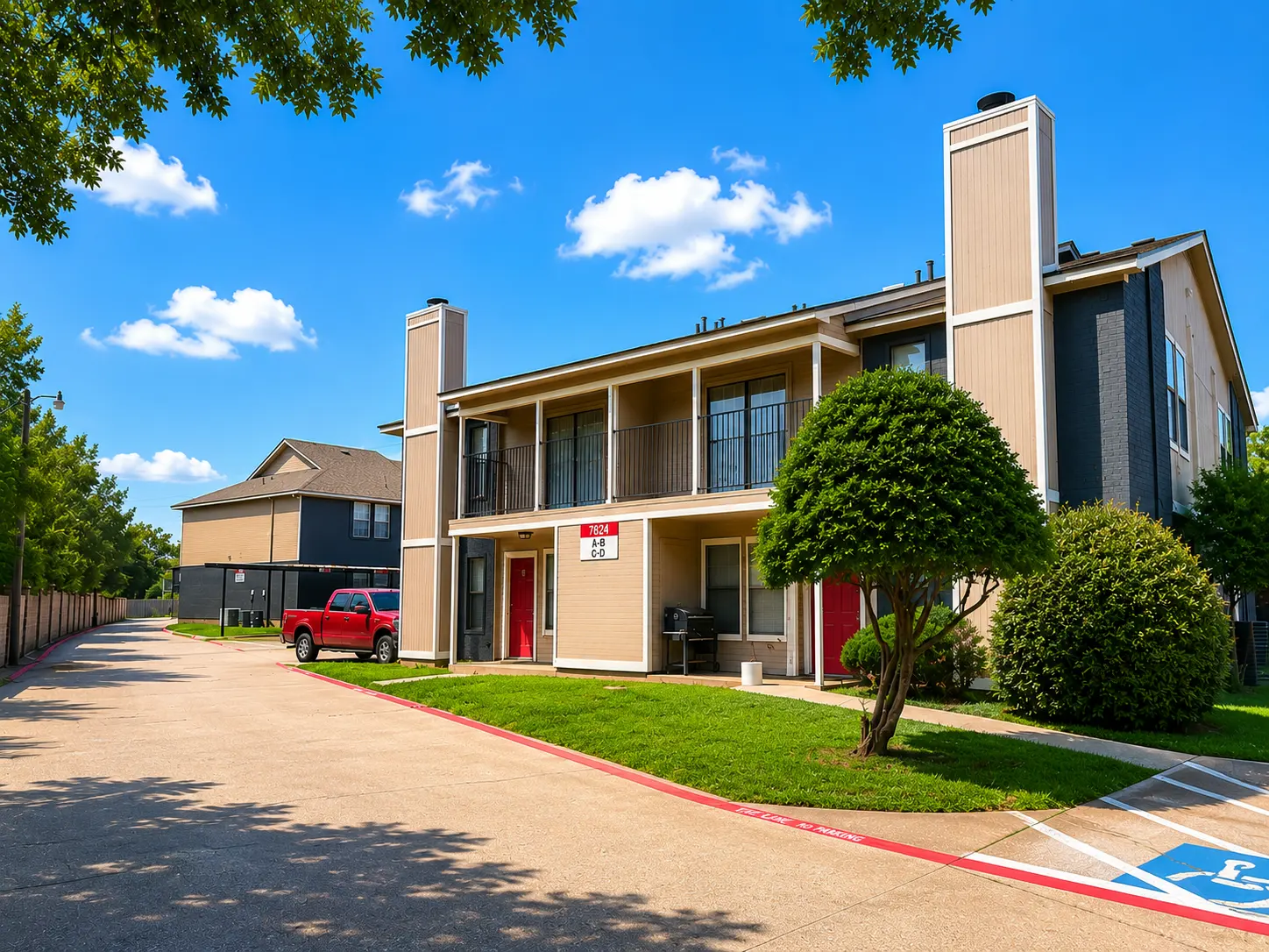 A view of multi-unit residential buildings with a parking area and green landscaping under a blue sky with scattered clouds.