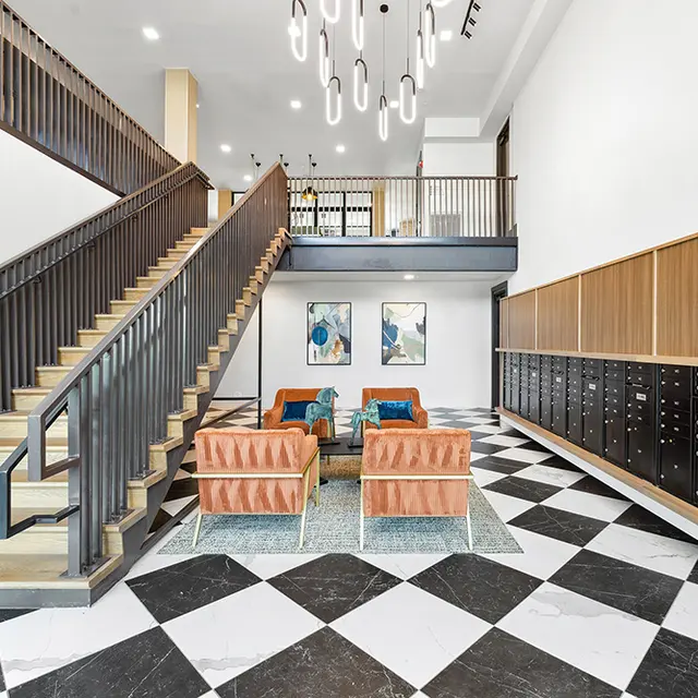 A spacious and modern lobby featuring a striking staircase, contemporary light fixtures, and stylish seating. The floor has a black and white checkered pattern, and there are wooden cubby storage units along the wall.