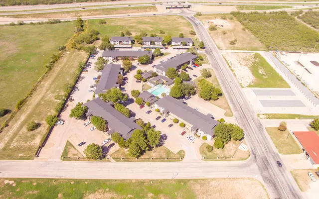 Aerial view of a sprawling apartment complex with multiple buildings arranged around a central pool area. Surrounding greenery and roads visible.