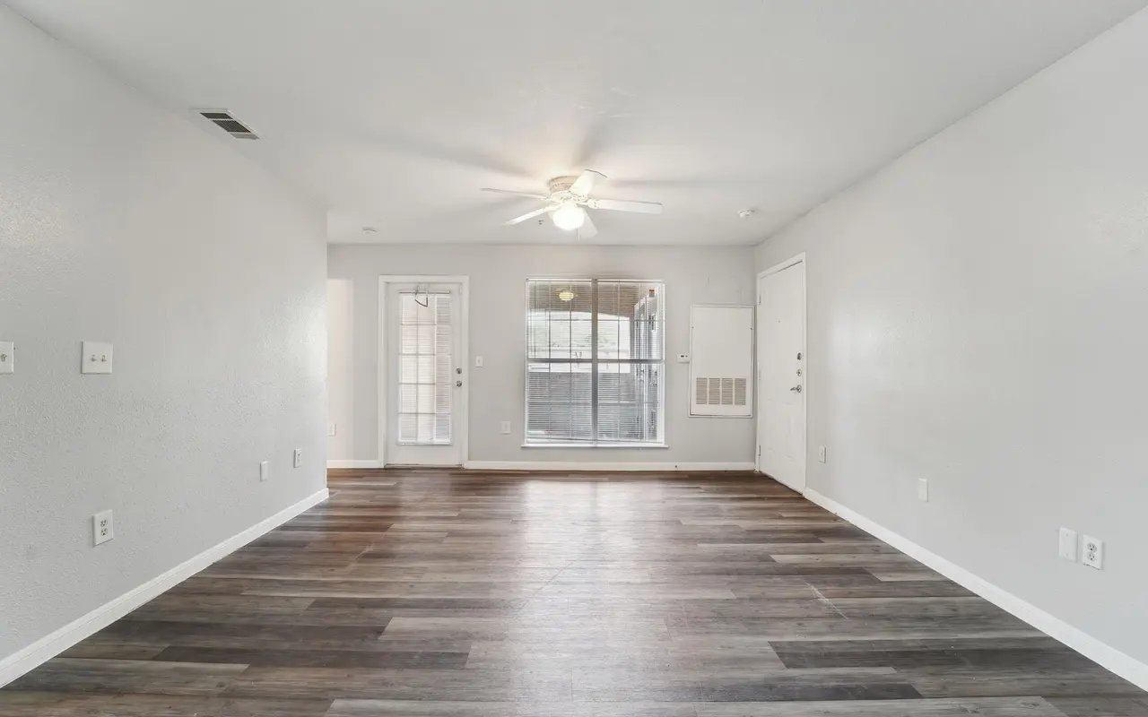 An empty living room with gray walls and wooden flooring. A ceiling fan is visible, and there are two doors leading to a balcony or patio area. There are light switches and outlets on the walls.