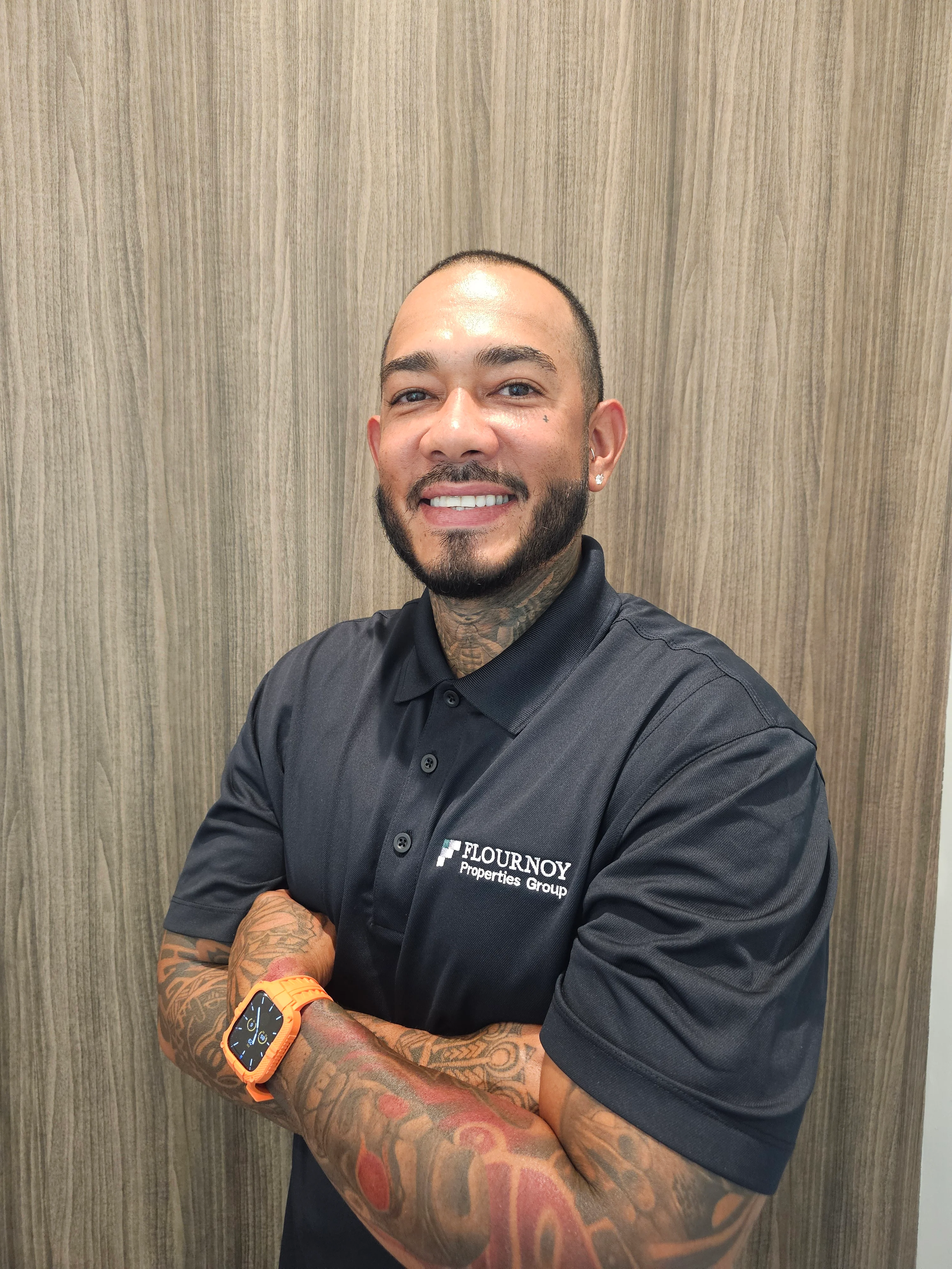Flournoy Properties Group Staff Member A smiling individual wearing a dark polo shirt with the logo of Flournoy Properties Group, standing against a wooden background, arms crossed, showcasing tattoos on the arms.