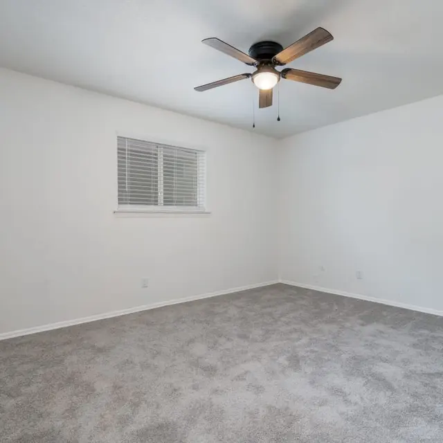 An empty bedroom featuring light gray carpet, a ceiling fan with wooden blades, and a window with white slats. The walls are painted white, providing a bright and spacious feel.