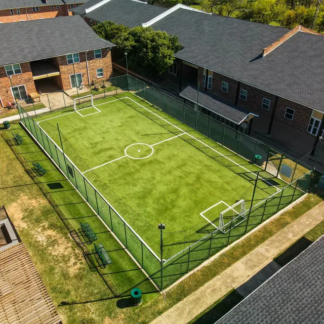 Aerial view of a small soccer field surrounded by residential buildings. The field has artificial turf, goal posts on each end, and a chain-link fence encircling it. Green benches are placed along the perimeter.