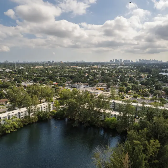 Aerial view of a lush green area with a lake surrounded by trees and residential buildings, under a partly cloudy sky.