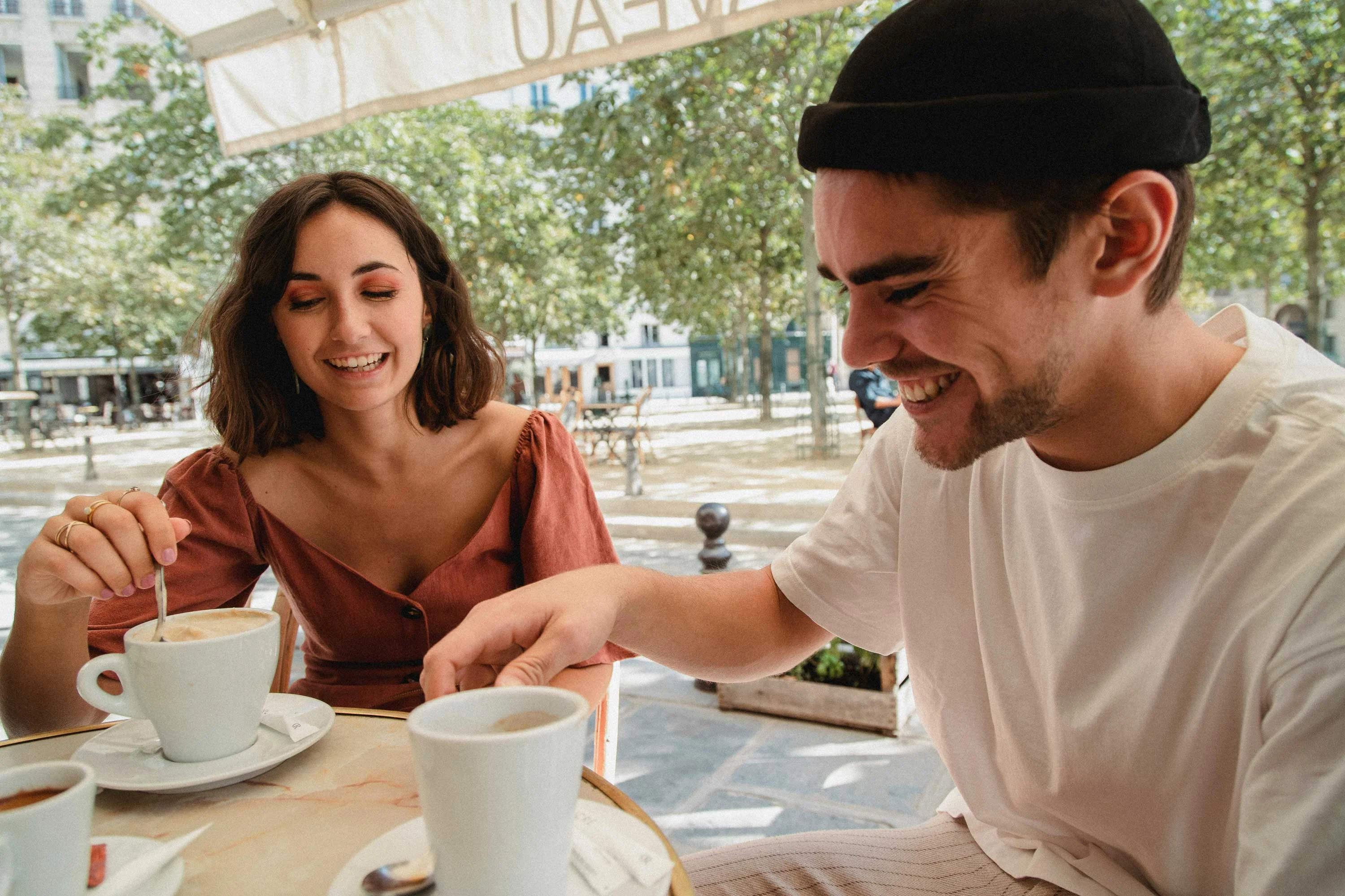 Joyful Coffee Date A man and woman enjoying coffee at a cafe, smiling and interacting in a sunny outdoor setting with green trees.