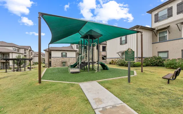 Residential Playground A playground with a green shade structure, surrounded by grass, and adjacent to residential buildings.
