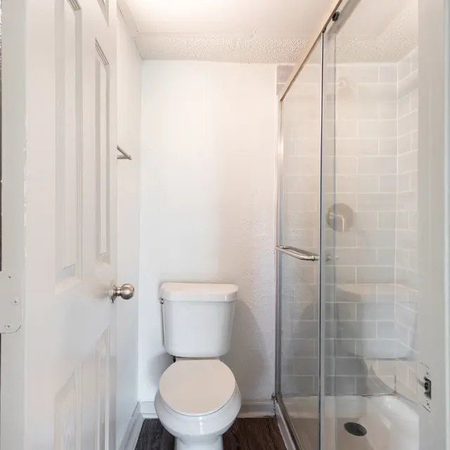 A modern bathroom featuring a white toilet and a glass shower enclosure. The walls are painted white, and the flooring is dark wood-like. A door is visible on the left side of the image.