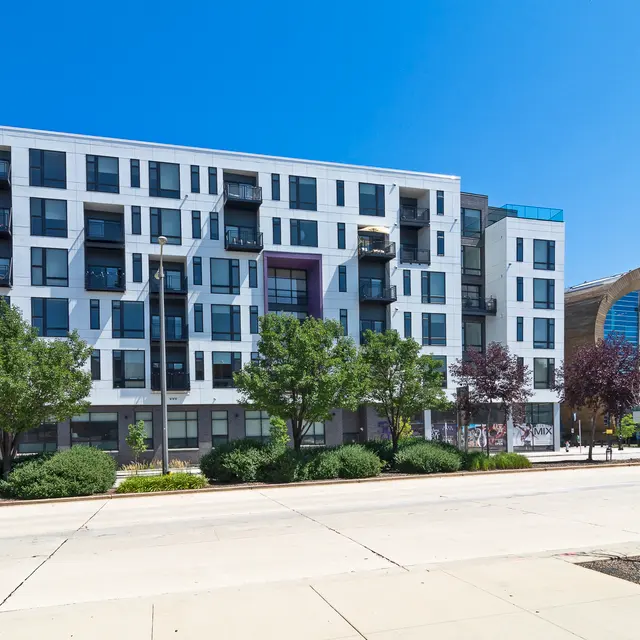 A modern apartment building with balconies, surrounded by trees, located next to a sports arena
