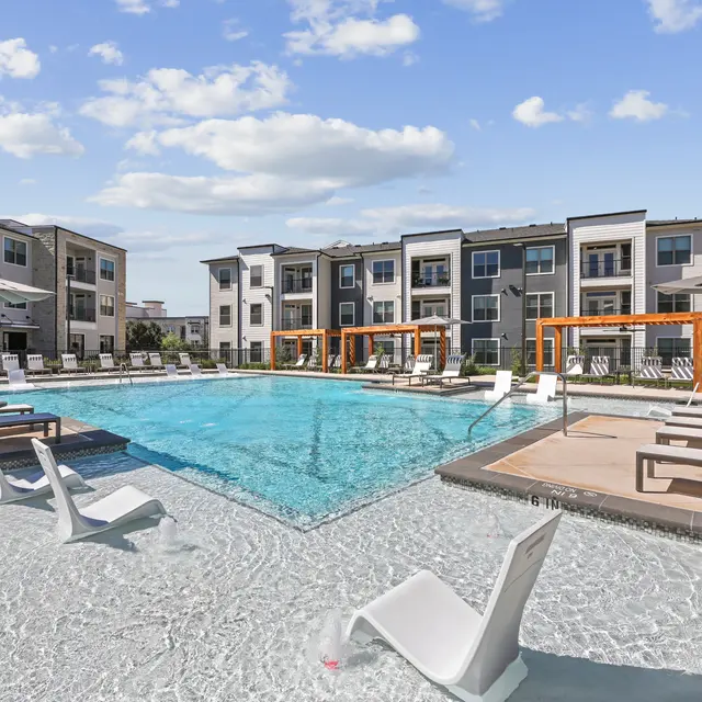 A modern apartment complex featuring a large swimming pool surrounded by lounge chairs and shaded seating areas.