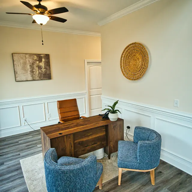 A modern office space featuring a wooden desk, two blue upholstered chairs, a ceiling fan, and light-colored walls with wainscoting. A small plant and a decorative wall piece add to the decor.