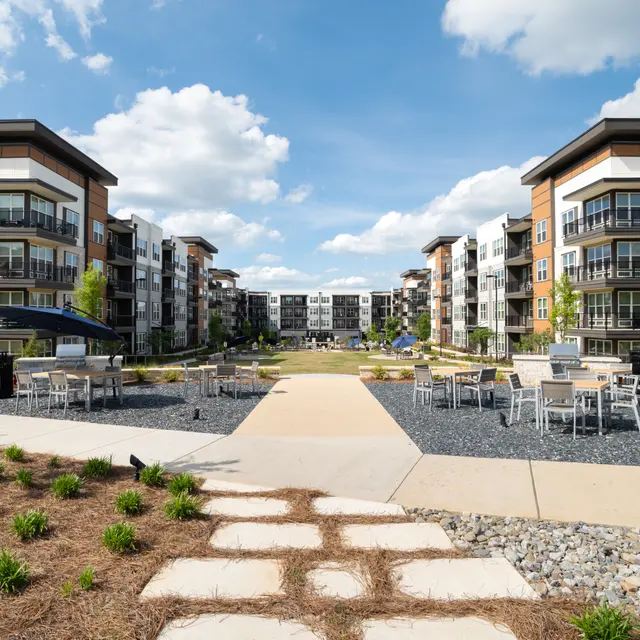 A view of a modern apartment complex featuring two multi-story buildings on either side of a landscaped pathway. The area includes outdoor seating and landscaping with grass and stones under a bright blue sky with scattered clouds.