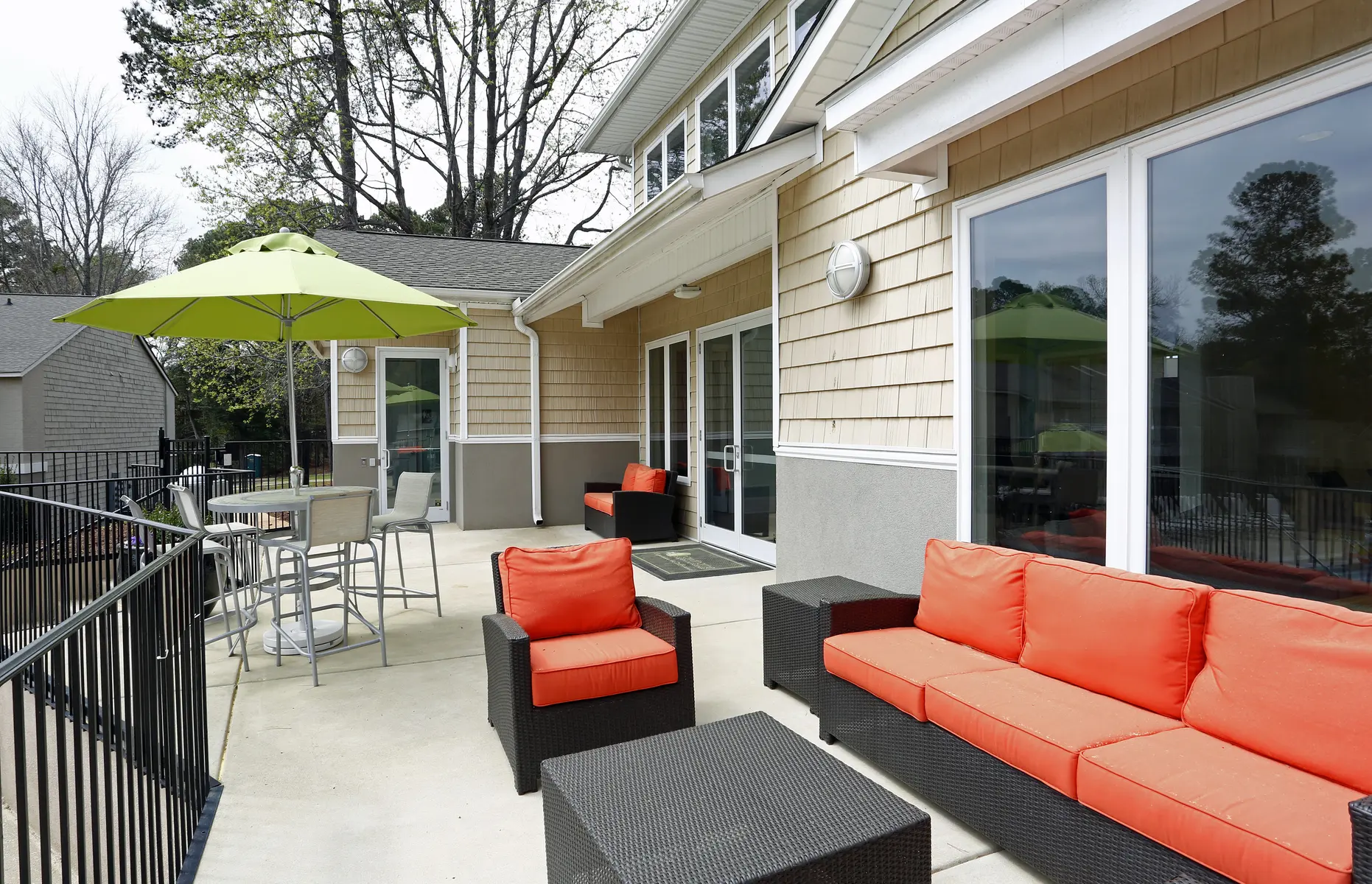 A modern outdoor lounge area with orange and black furniture, including a sectional sofa and a table with chairs. A green umbrella provides shade, and the setting has a mix of greenery and a cozy atmosphere.