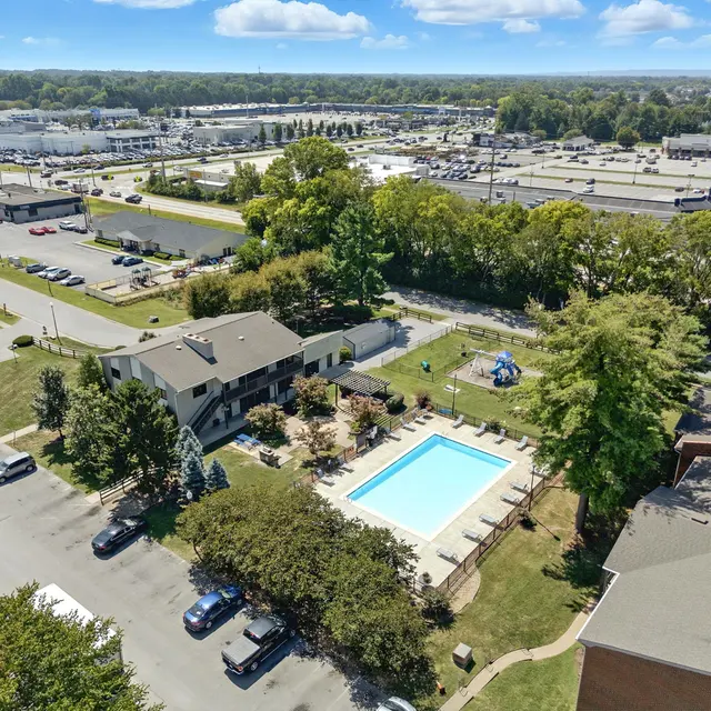 Aerial view of an apartment complex featuring a swimming pool and surrounding greenery, with parking lots and commercial buildings in the background.