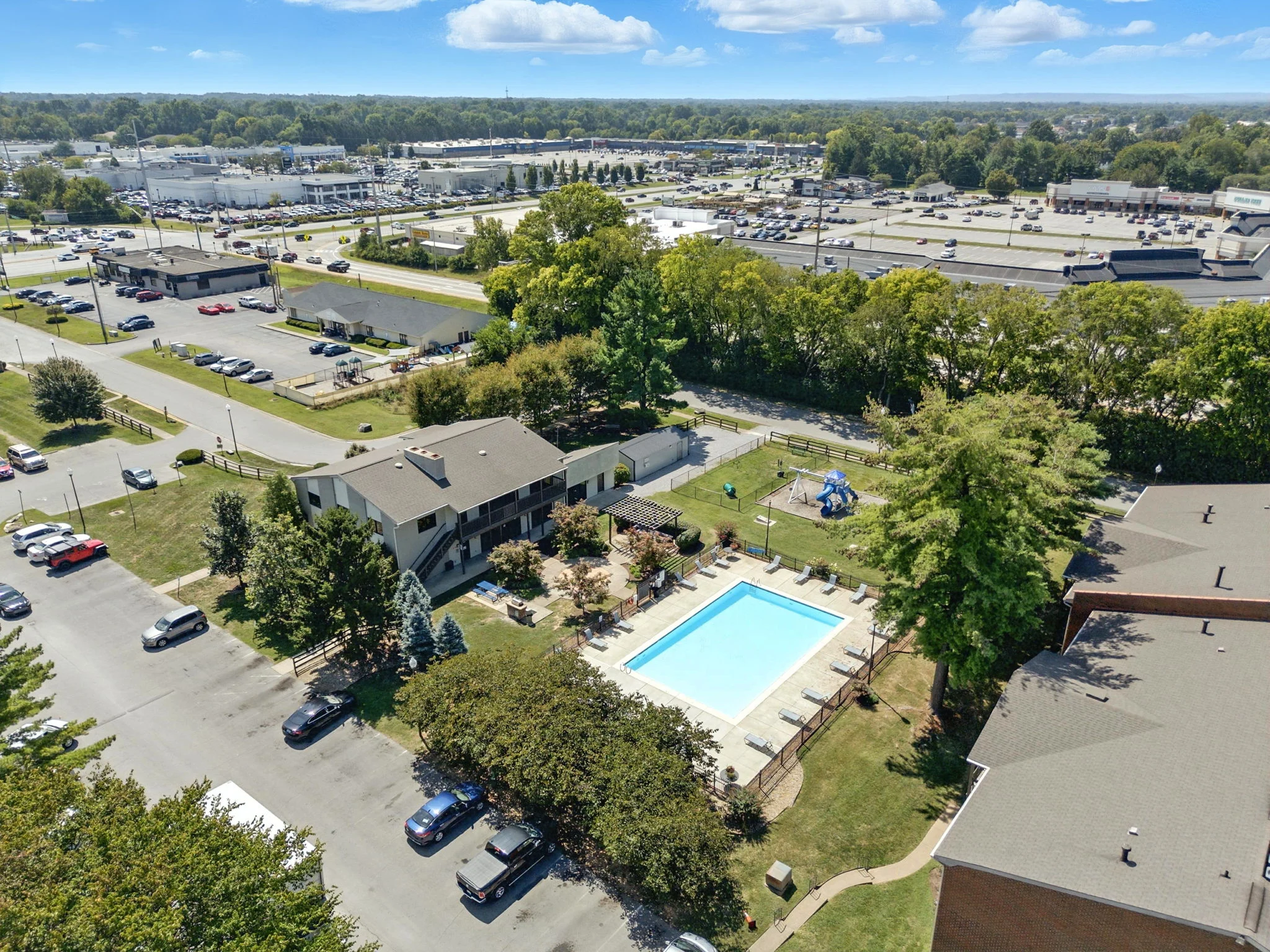 Aerial view of an apartment complex featuring a swimming pool and surrounding greenery, with parking lots and commercial buildings in the background.