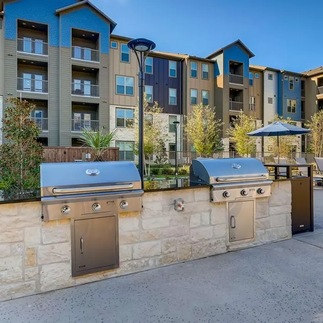 A modern apartment complex with a grilling area, featuring two stainless steel grills, shaded seating areas, and well-maintained landscaping.