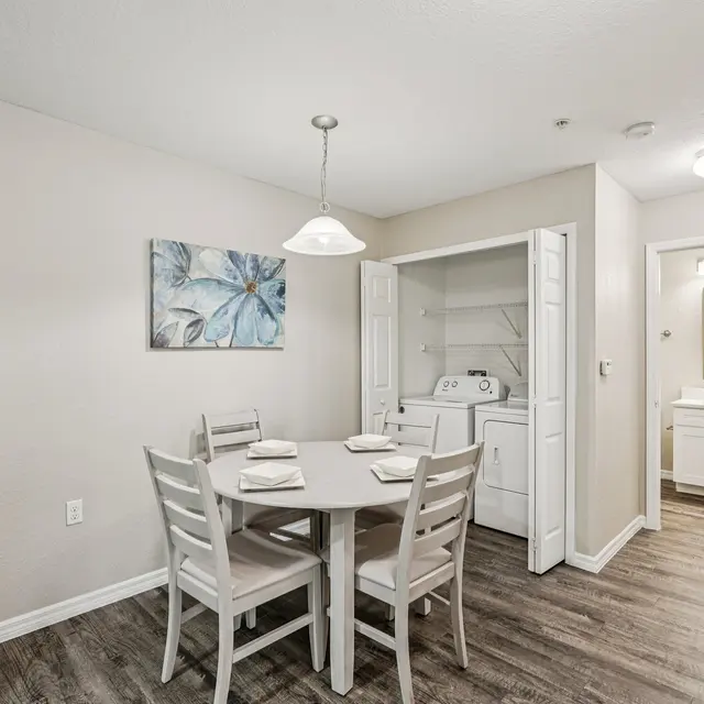 A small dining area featuring a round table with four chairs, a decorative painting on the wall, and a laundry closet in the background.