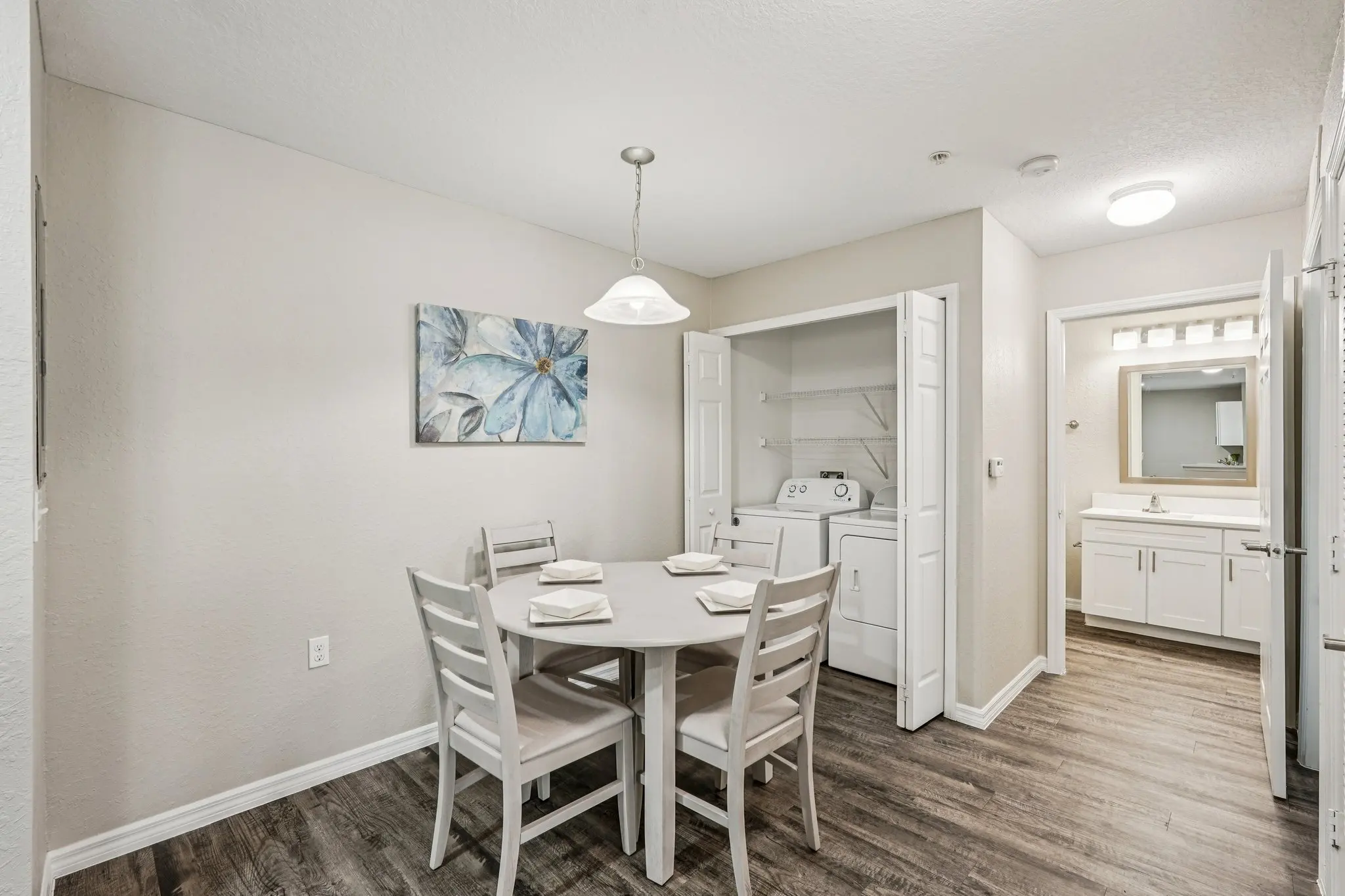 A small dining area featuring a round table with four chairs, a decorative painting on the wall, and a laundry closet in the background.