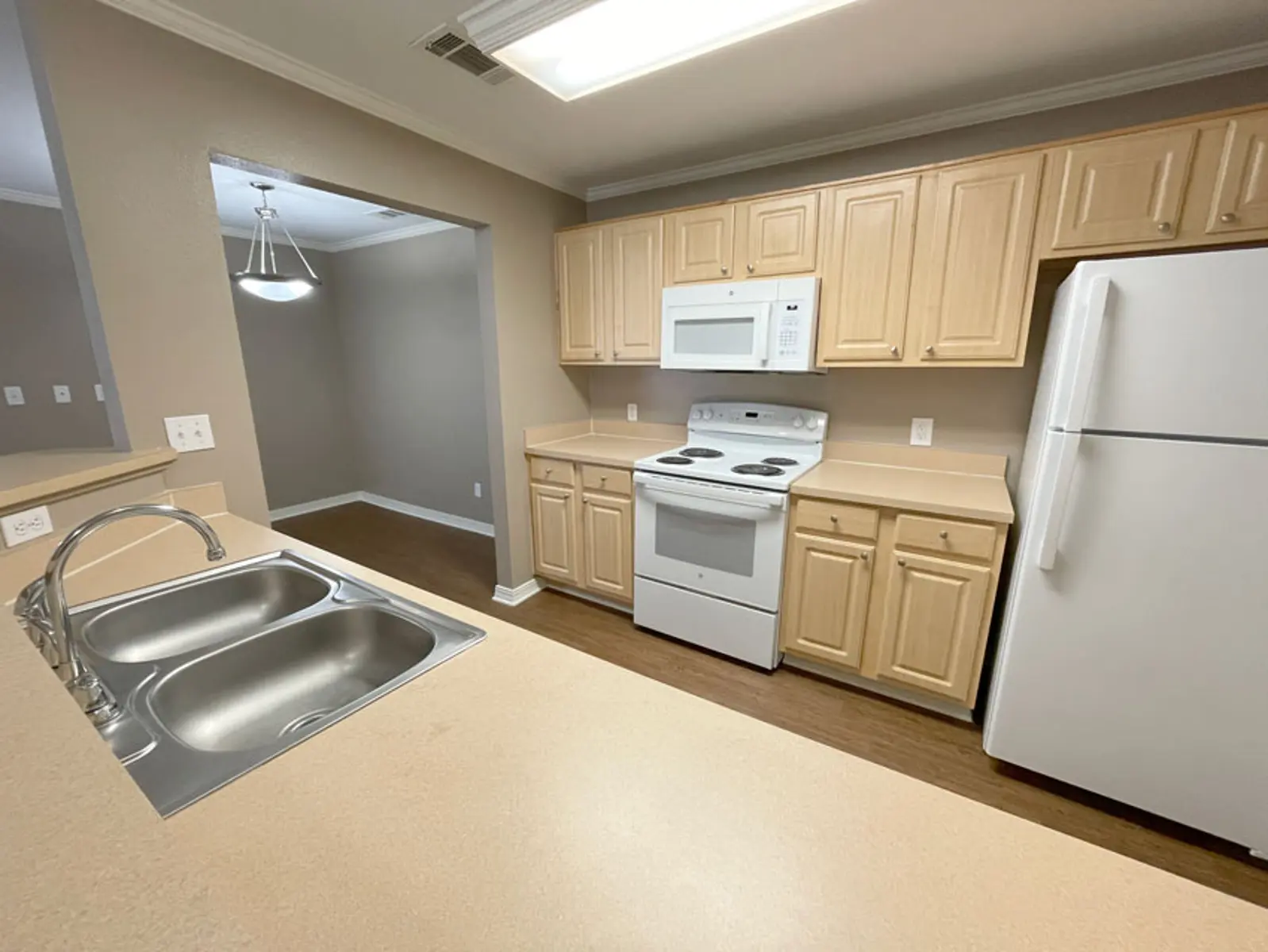 A modern kitchen with beige cabinetry, a white stove, microwave, and refrigerator, and a double sink. The space features a light brown floor and an open layout with a dining area visible in the background.