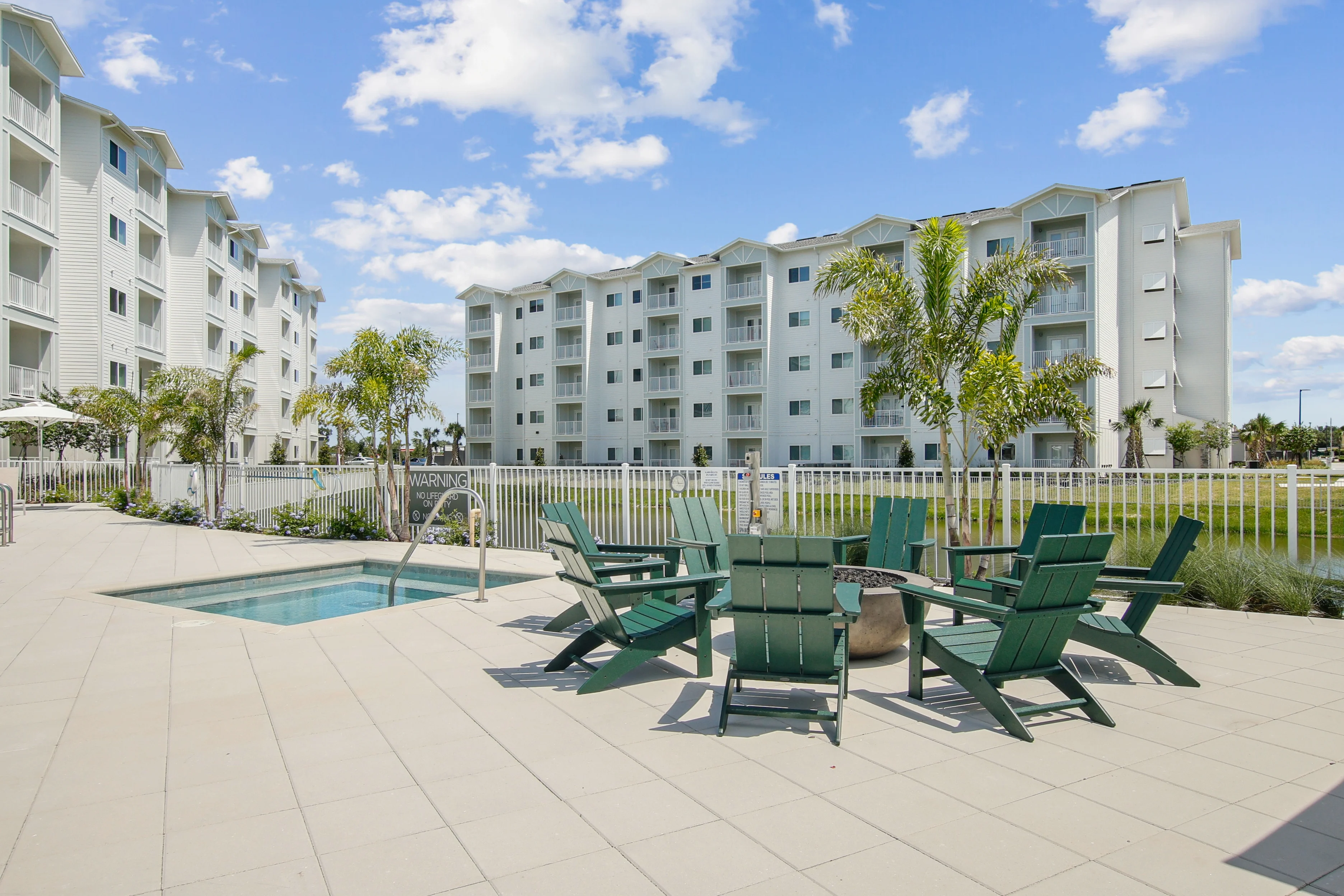 Sundance Trails A modern residential area featuring a small pool, green chairs, and buildings under a blue sky with fluffy clouds.