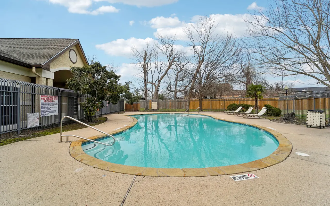 A tranquil outdoor swimming pool surrounded by a concrete deck and a fenced area. The pool has clear blue water and a gentle slope entry. There are a few lounge chairs positioned around the pool, and the background features bare trees and a wooden fence under a cloudy sky.