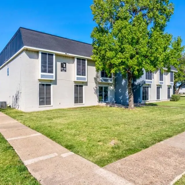 Exterior view of a two-story apartment building with green grass and trees around it, under a clear blue sky.