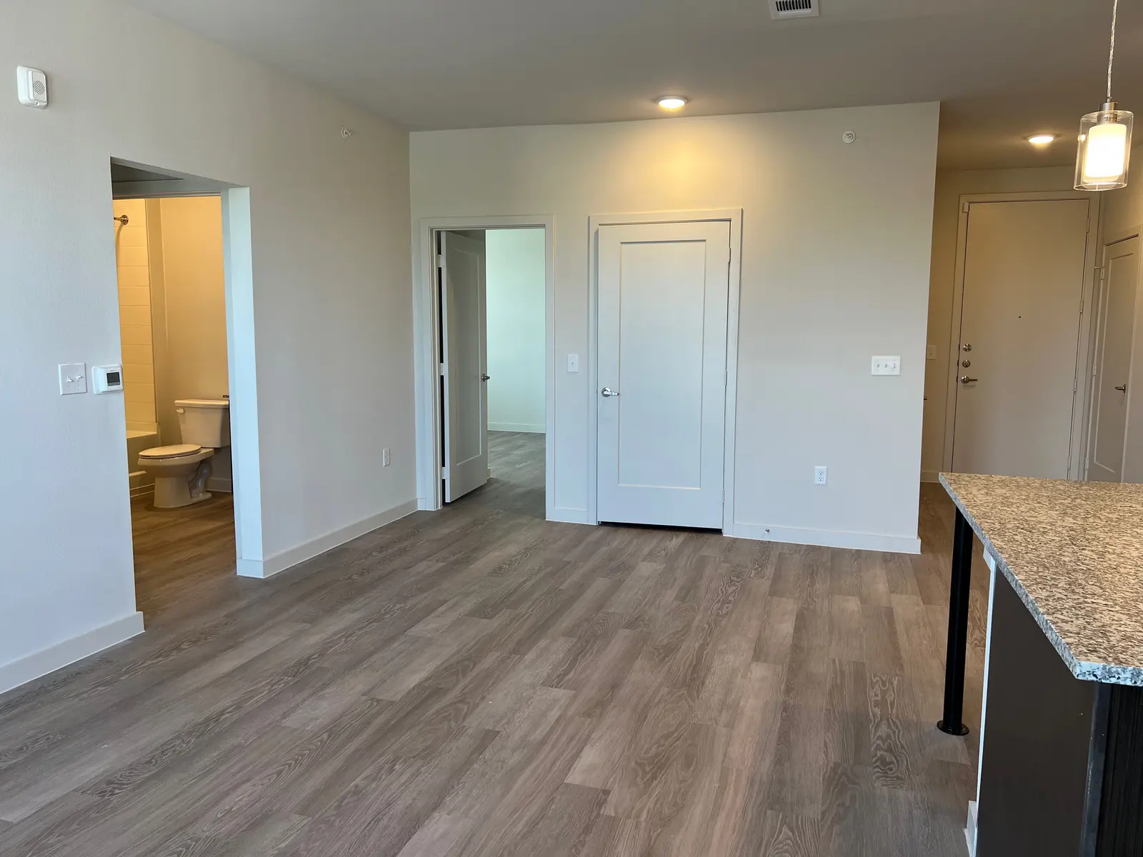 A spacious, modern apartment interior with light-colored walls and hardwood floors. The room features a kitchen area with a granite countertop on the right, and doorways leading to other rooms in the background. There is a bathroom visible through an open door on the left side.