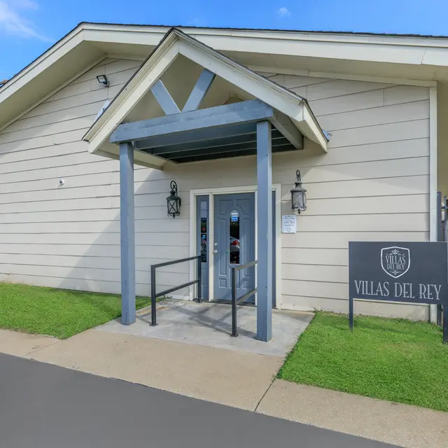Villas Del Rey Entrance Exterior view of a building labeled 'Villas Del Rey'. The entrance features a covered porch with a blue ceiling and decorative columns. Grass and a paved walkway are visible in the foreground.