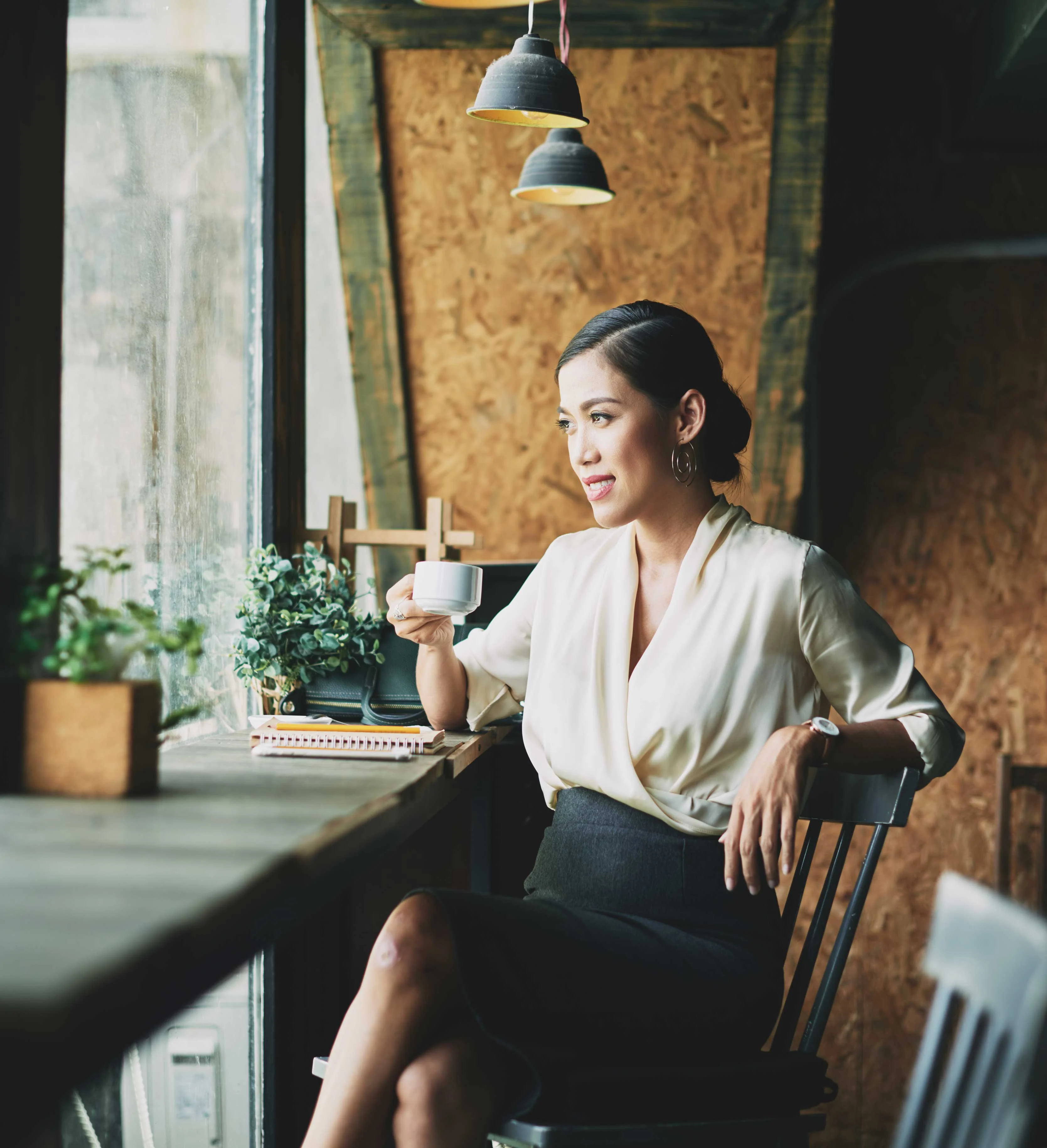 A woman sitting in a cafe, holding a coffee cup, dressed in a stylish blouse and skirt. She has a thoughtful expression, with natural light coming through the window, and plants arranged on the table nearby.