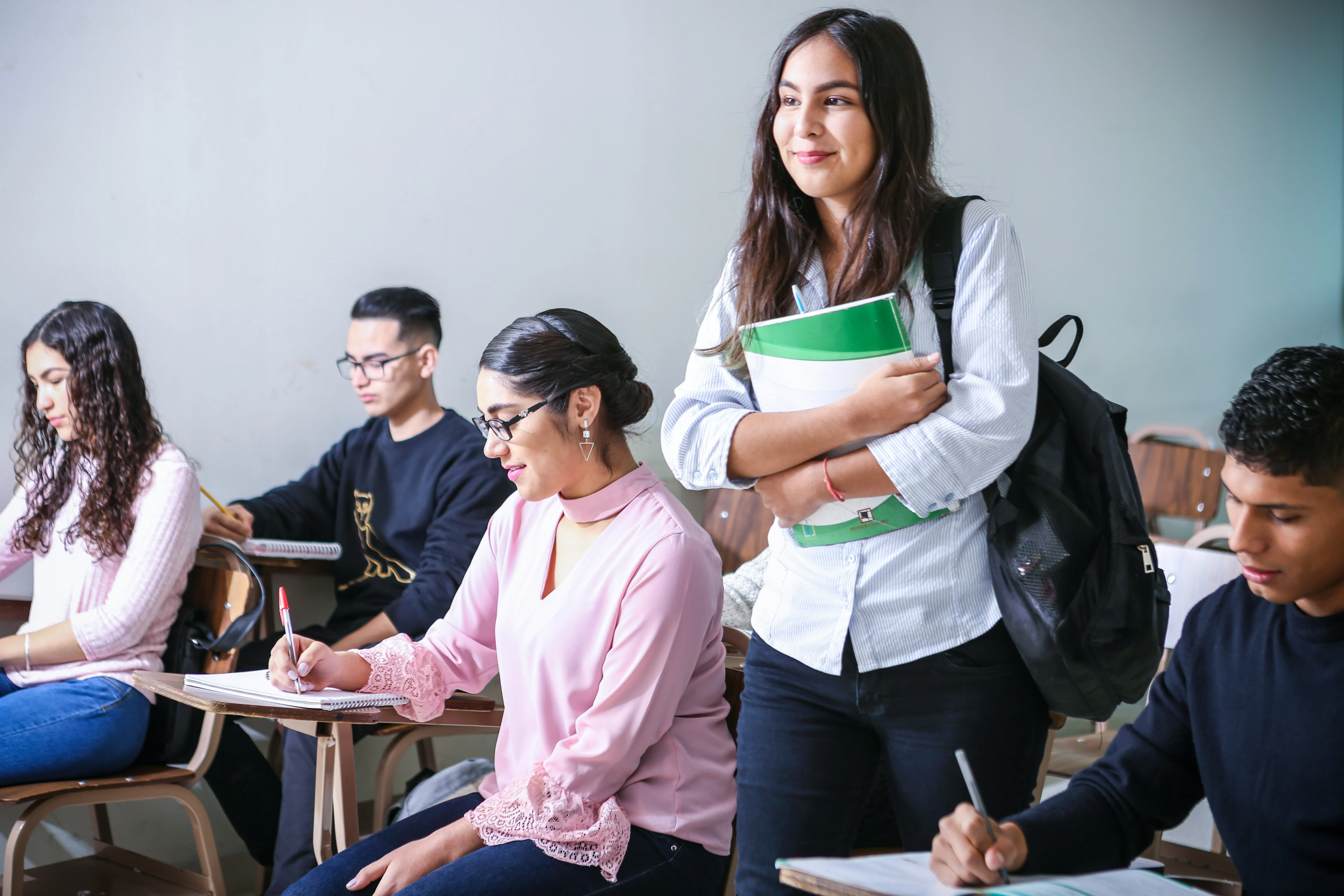 Classroom Dynamics A group of students in a classroom setting. Some are writing at their desks while one girl stands, holding books and looking at a classmate.