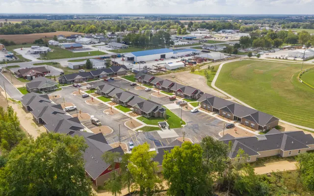 Aerial view of a neighborhood featuring several single-story homes arranged in a circular layout, surrounded by trees and a clear blue sky. In the background, there are additional buildings and open fields.