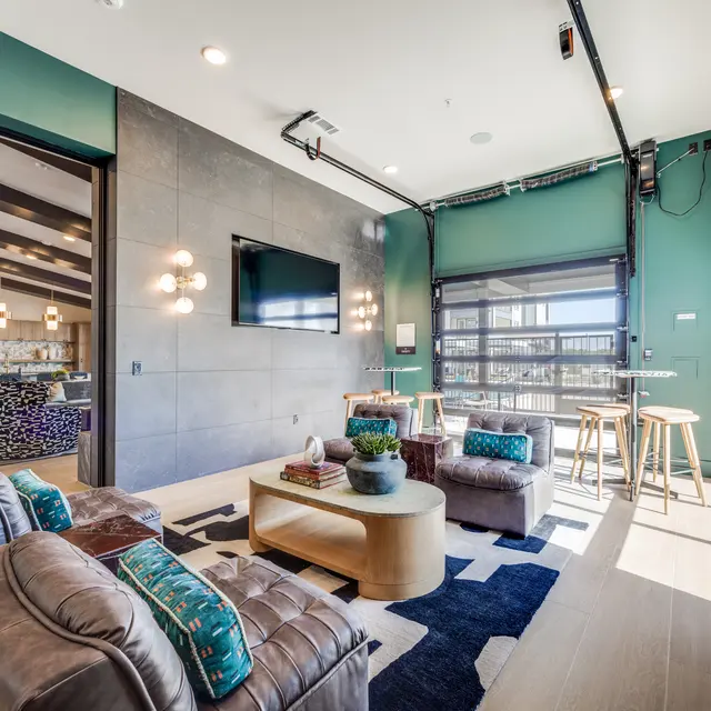A modern living space featuring a cozy seating area with brown sofas and a coffee table, large windows, and a garage door that opens to a view. The walls are painted an emerald green and there are bar stools and a flat-screen TV mounted on the wall.