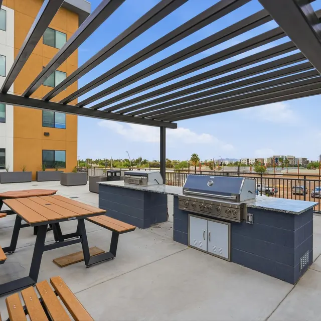 Outdoor Grilling Area An outdoor grilling area featuring a large stone grill station and wooden picnic tables shaded by a metal pergola against a modern residential building.