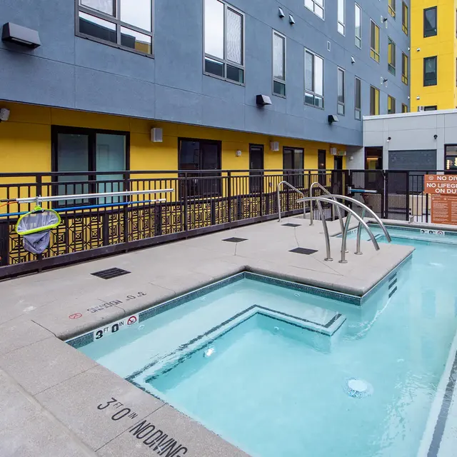 A modern pool area featuring a small hot tub surrounded by a tiled deck and a black railing. The background shows a building with yellow and gray exterior. A life preserver is visible on the railing.