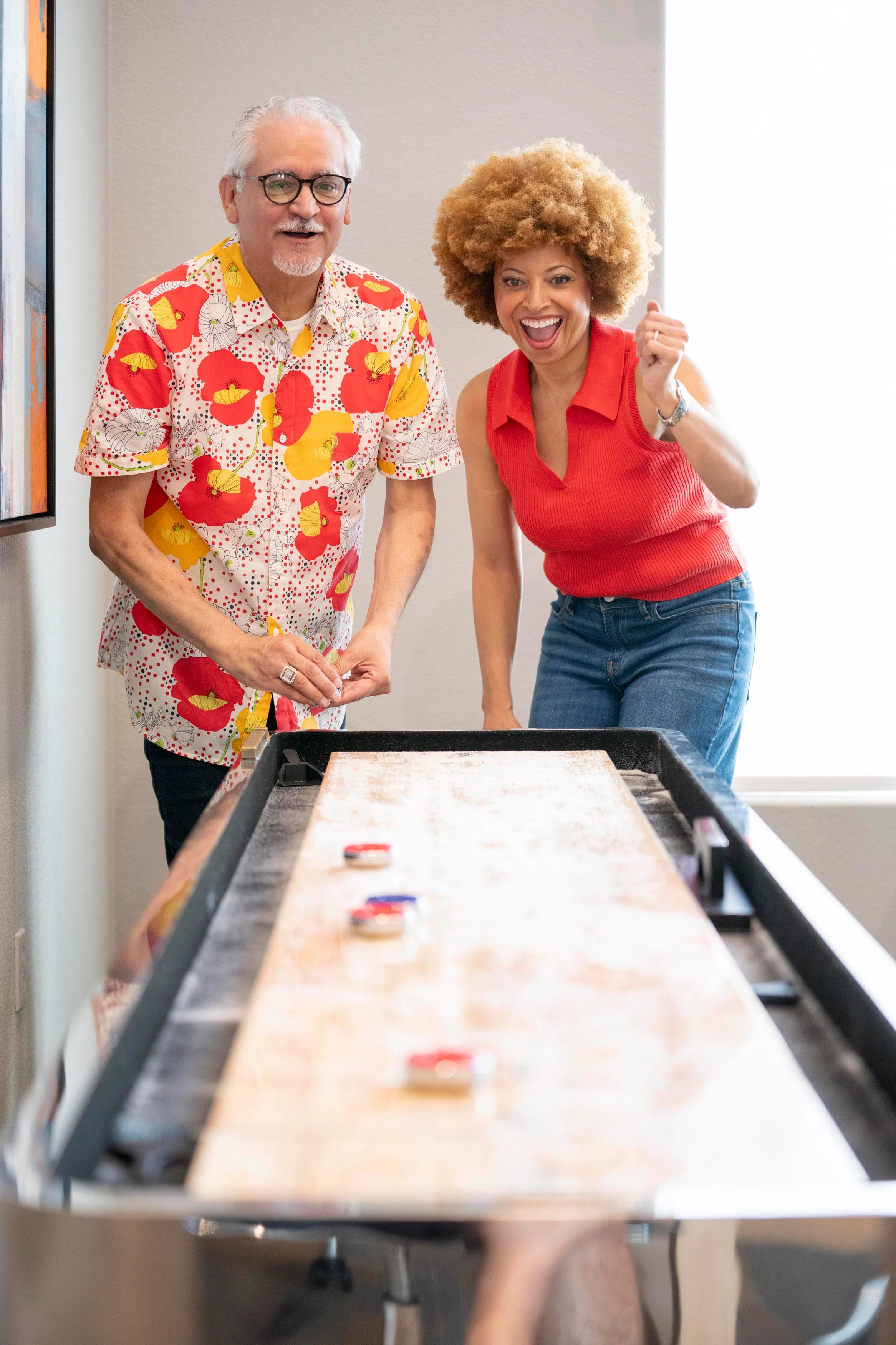 Two people enjoying a game of shuffleboard in a bright, modern interior. One person is wearing a colorful floral shirt, while the other is dressed in a red top and denim pants. They both appear to be in a playful, competitive mood.
