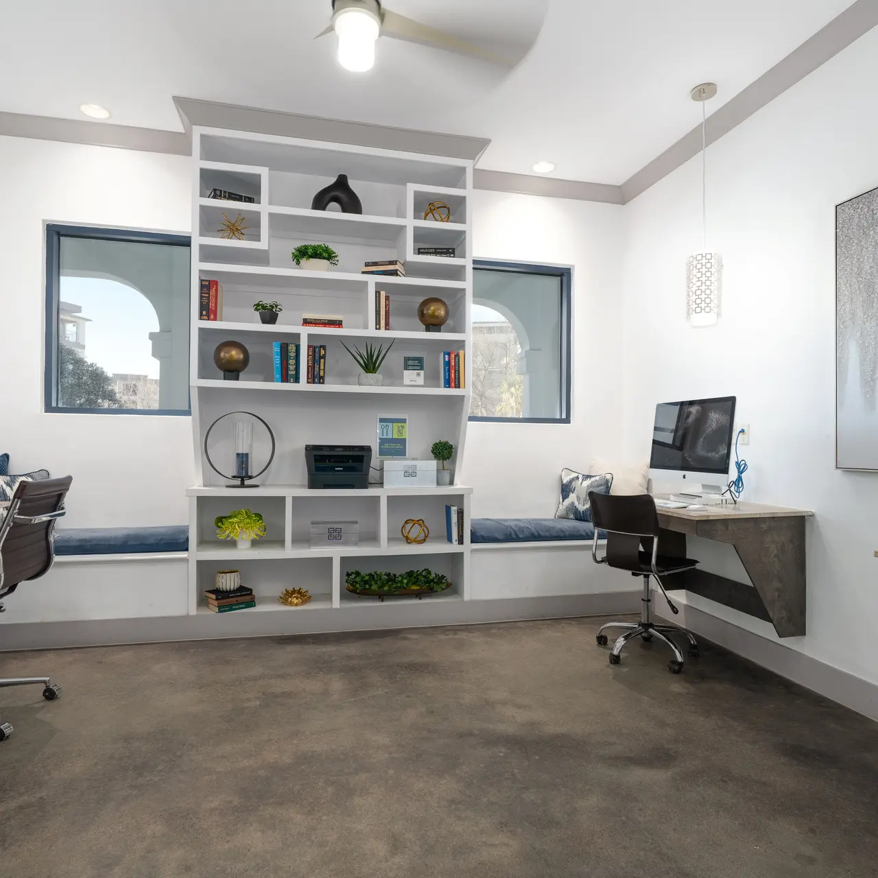 A modern home office featuring two workstations, a built-in bookshelf, and large windows allowing natural light to illuminate the space. The floor is concrete, and there are decorative elements on the shelves and tables.