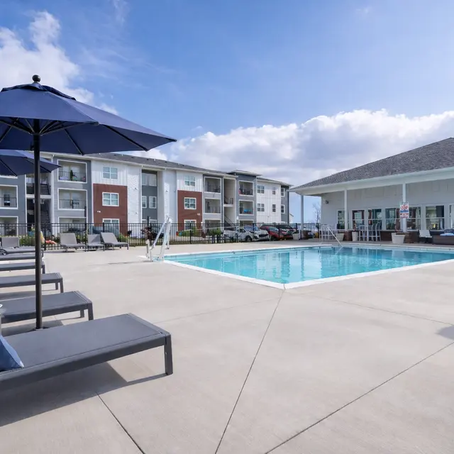 Swimming pool area at a luxury apartment complex featuring lounge chairs and umbrellas, surrounded by landscaped grounds.