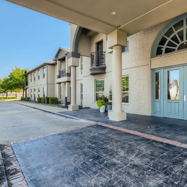 View of a hotel entrance with large double doors and a patio area. The surrounding landscape includes a paved pathway and some greenery.