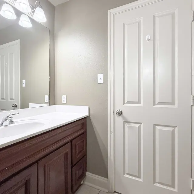 A modern bathroom featuring a white countertop sink with two faucets, a large mirror above it, and a brown wooden cabinet beneath. The wall is painted in a light shade, and there is a closed white door on the right side.