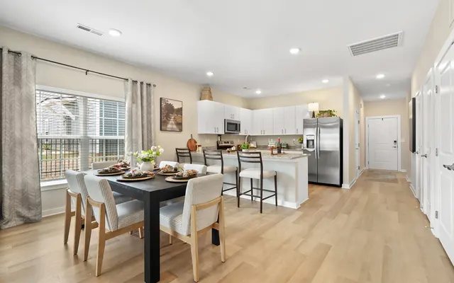 A modern kitchen and dining area featuring a small dining table set with dishes, surrounded by chairs. The kitchen includes white cabinets, stainless steel appliances, and a window allowing natural light.