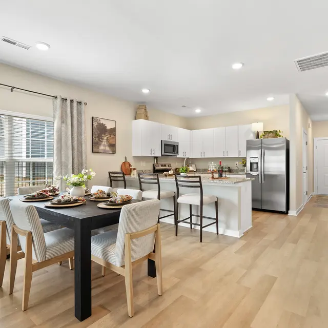 A modern kitchen and dining area featuring a small dining table set with dishes, surrounded by chairs. The kitchen includes white cabinets, stainless steel appliances, and a window allowing natural light.