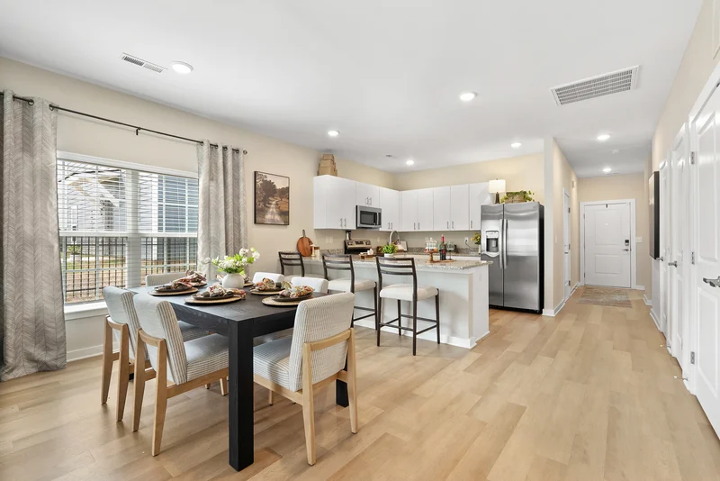 A modern kitchen and dining area featuring a small dining table set with dishes, surrounded by chairs. The kitchen includes white cabinets, stainless steel appliances, and a window allowing natural light.