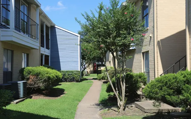 A sunny view of a well-maintained apartment complex courtyard, featuring a narrow pathway lined with shrubs and small trees, connecting two buildings with balconies.