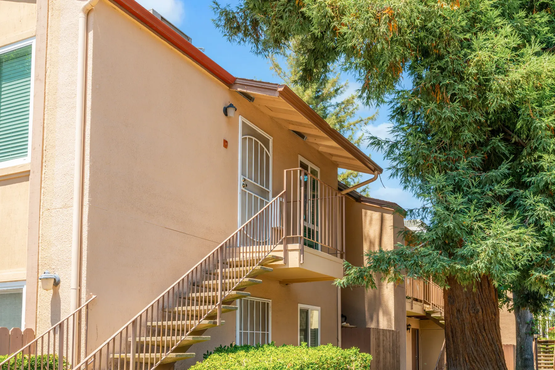 A light-colored two-story apartment building with a staircase leading to the upper floor, surrounded by greenery and tall trees.