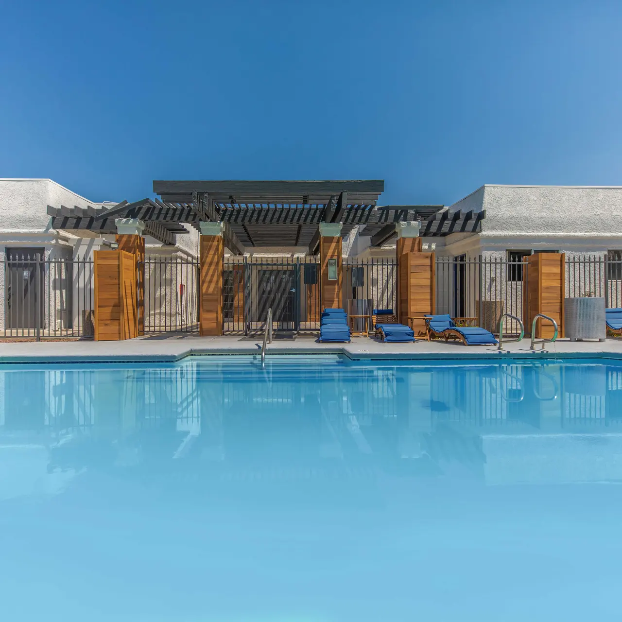 A clear blue swimming pool surrounded by lounge chairs and wooden structures, under a bright blue sky.