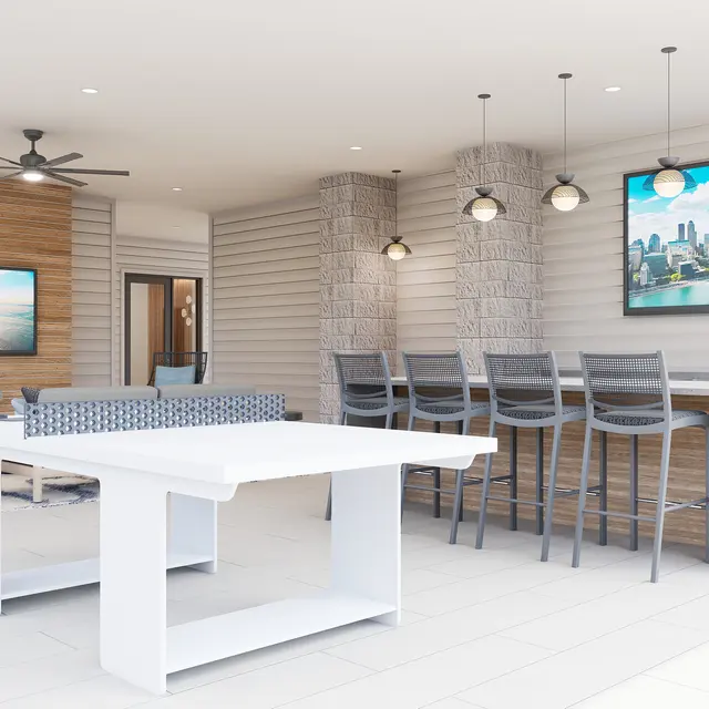 A modern and spacious indoor living area featuring a large white table, bar-style seating, and two televisions mounted on the walls. The decor includes wooden accents and a light color palette with large windows allowing natural light.