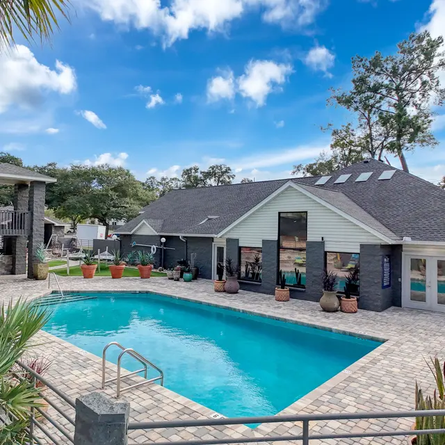 A spacious swimming pool area featuring a clear pool surrounded by vibrant greenery and potted plants, with a modern building in the background.